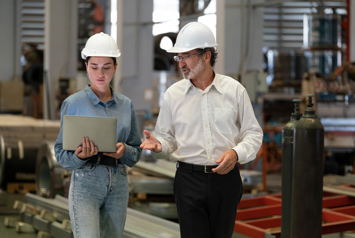 Eine Frau und ein Mann mit weißen Schutzhelmen gehen durch einen industriellen Fertigungsbereich, während sie Informationen auf einem Laptop überprüfen