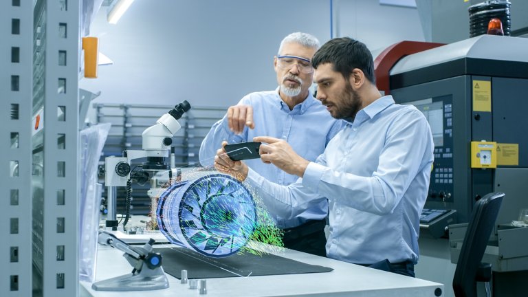 Dois engenheiros em uma fábrica observando um celular. Há uma peça sobre a mesa 