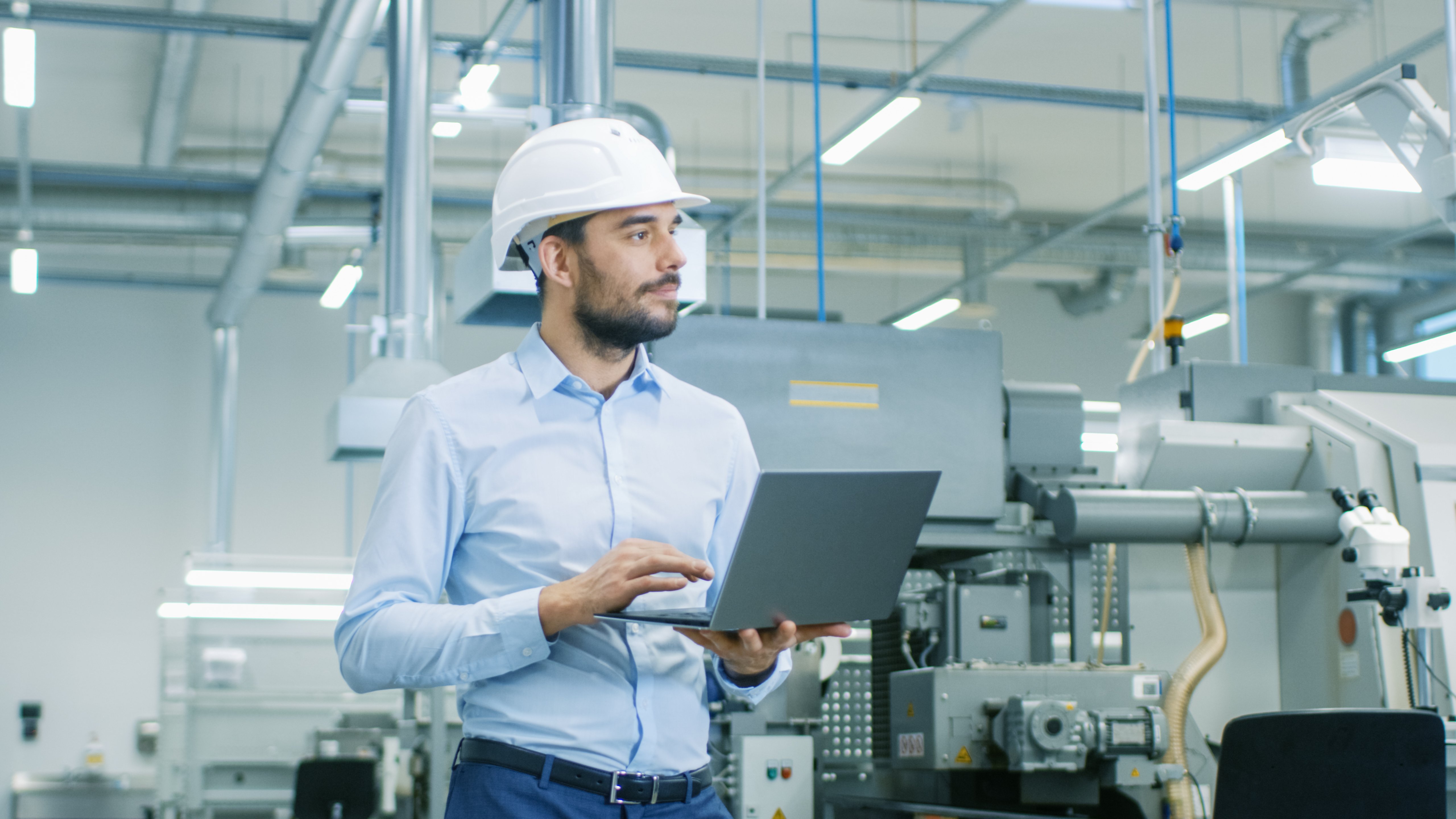 Ein Ingenieur steht in einem Werk, schaut sich laufende Maschinen an und tippt Informationen in seinen Laptop ein