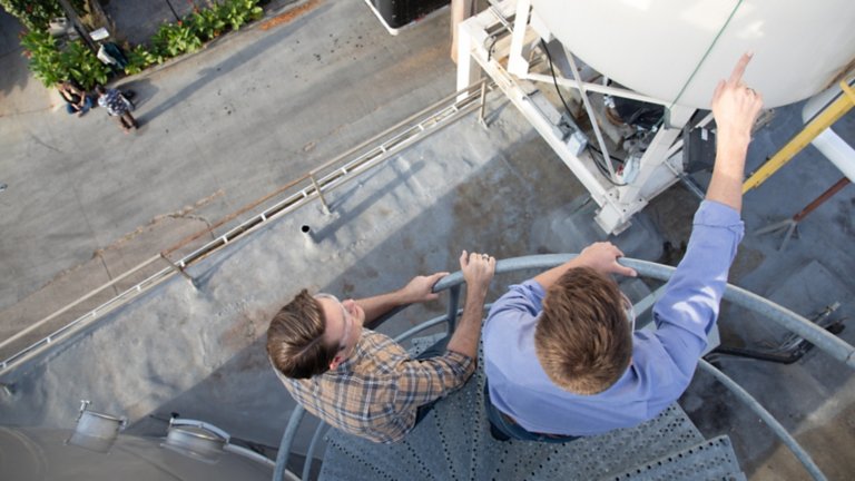 Dois engenheiros na parte externa das instalações, em pé no alto de uma escada caracol, apontando para um tanque contendo líquido.