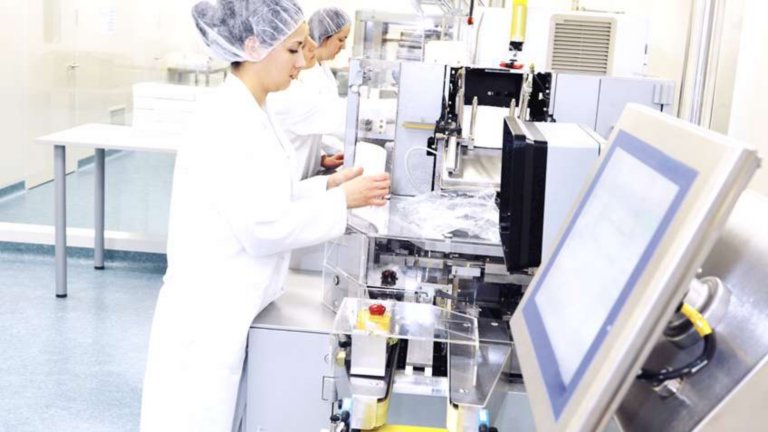 Two female life sciences workers on a pharmaceutical production line perform tasks at their stations. 