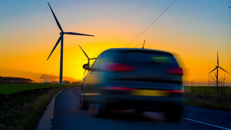 Auto, das bei Sonnenunter- oder -aufgang an einem klaren Wintertag auf einer Landstraße im Vereinigten Königreich an Feldern mit Windkraftanlagen vorbeifährt