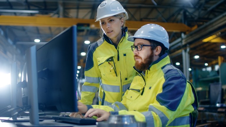 Dois funcionários vendo um monitor em uma fábrica usando jaquetas de segurança amarelas e capacetes brancos.