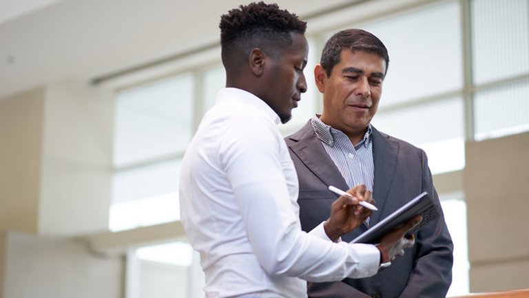 Dois homens conversando em ambiente de escritórios, um deles está segurando um tablet e uma caneta