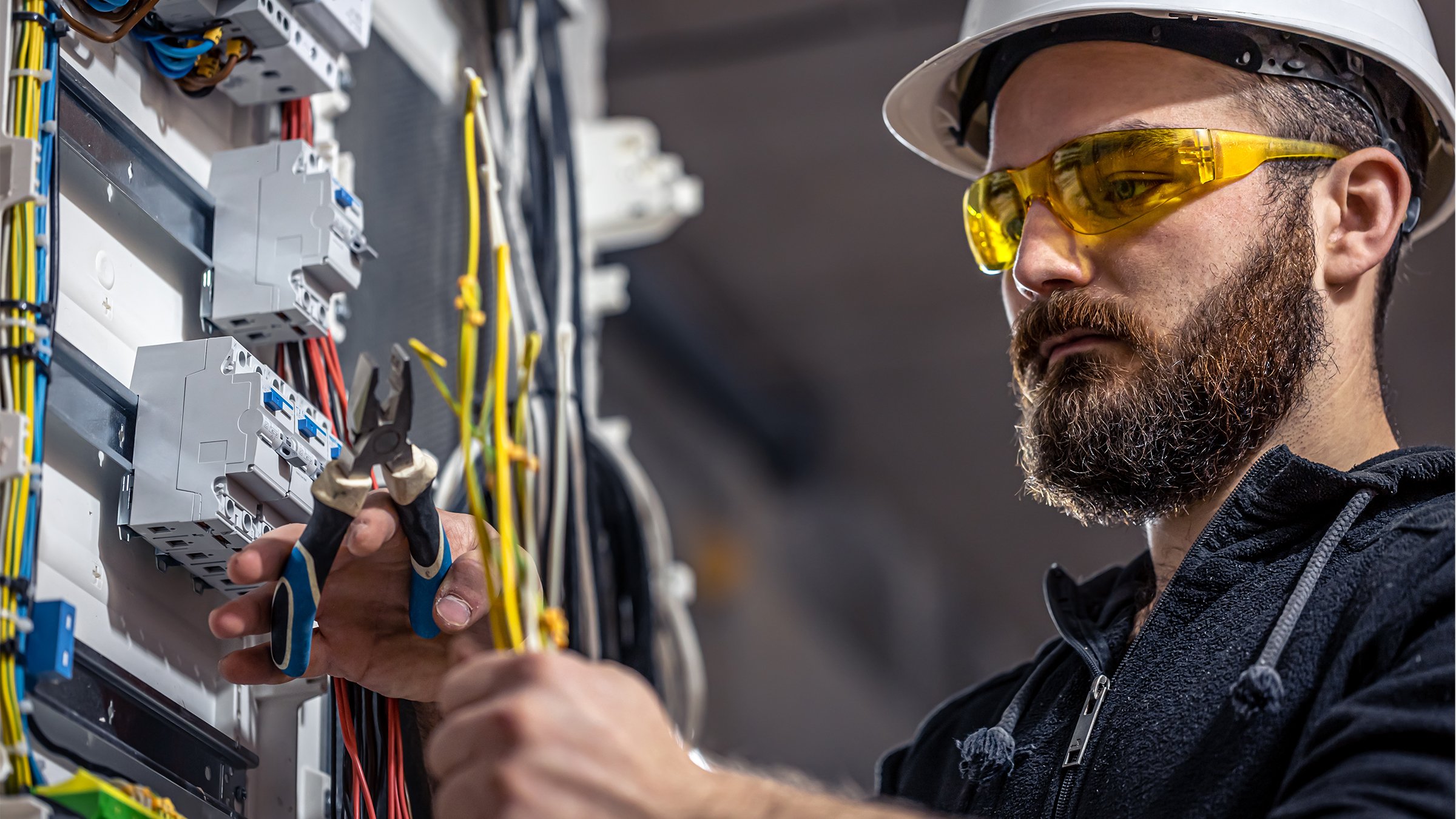 A skilled electrician in a hard hat and safety glasses carefully working with pliers on a complex electrical panel.