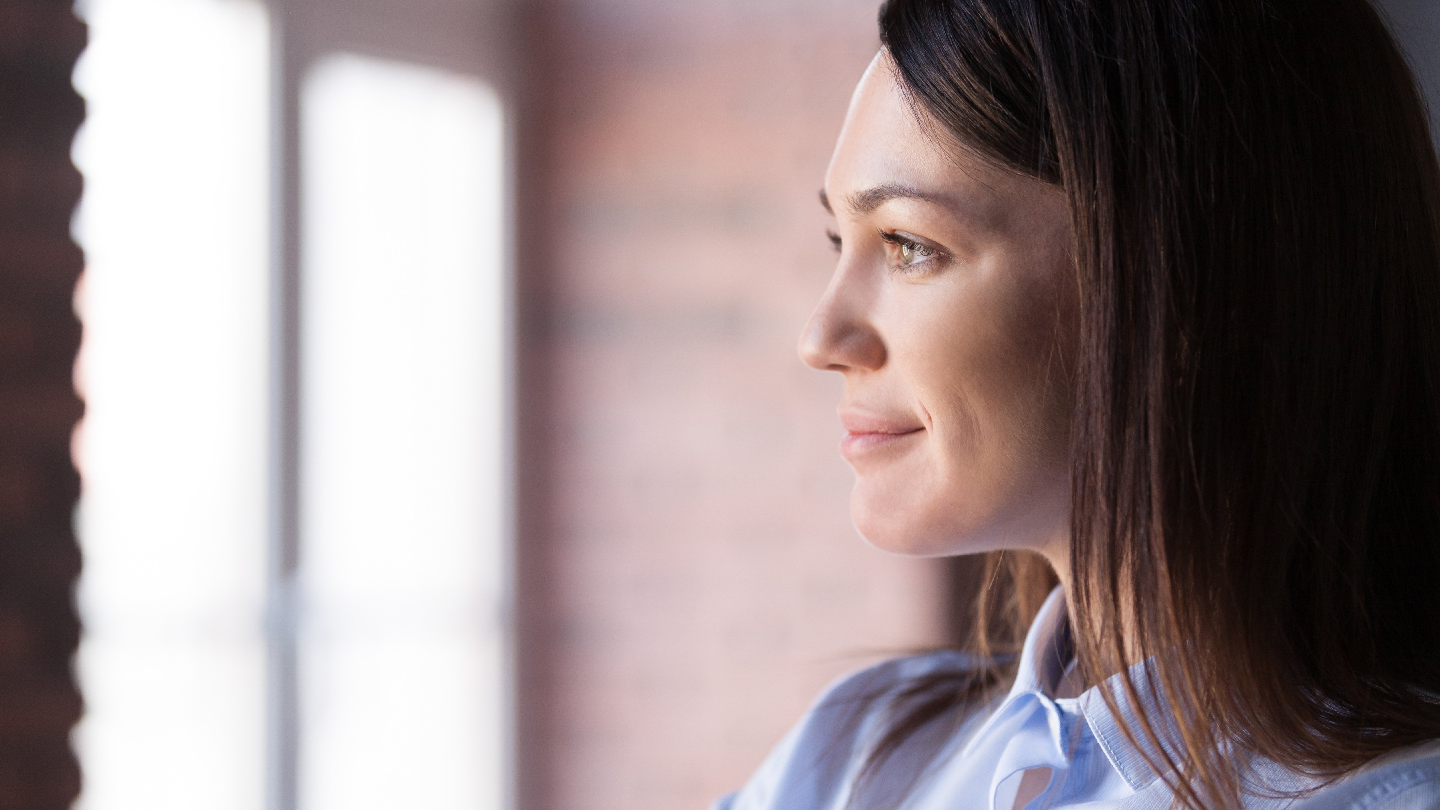 Female executive smiling with dark brown hair and wearing a light blue blouse and blazer looking out a window