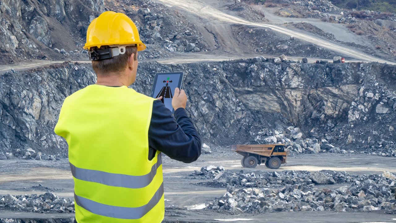 An engineer holds a tablet in his hand and remotely controls a heavy-duty dump truck in the background of a mining quarry
