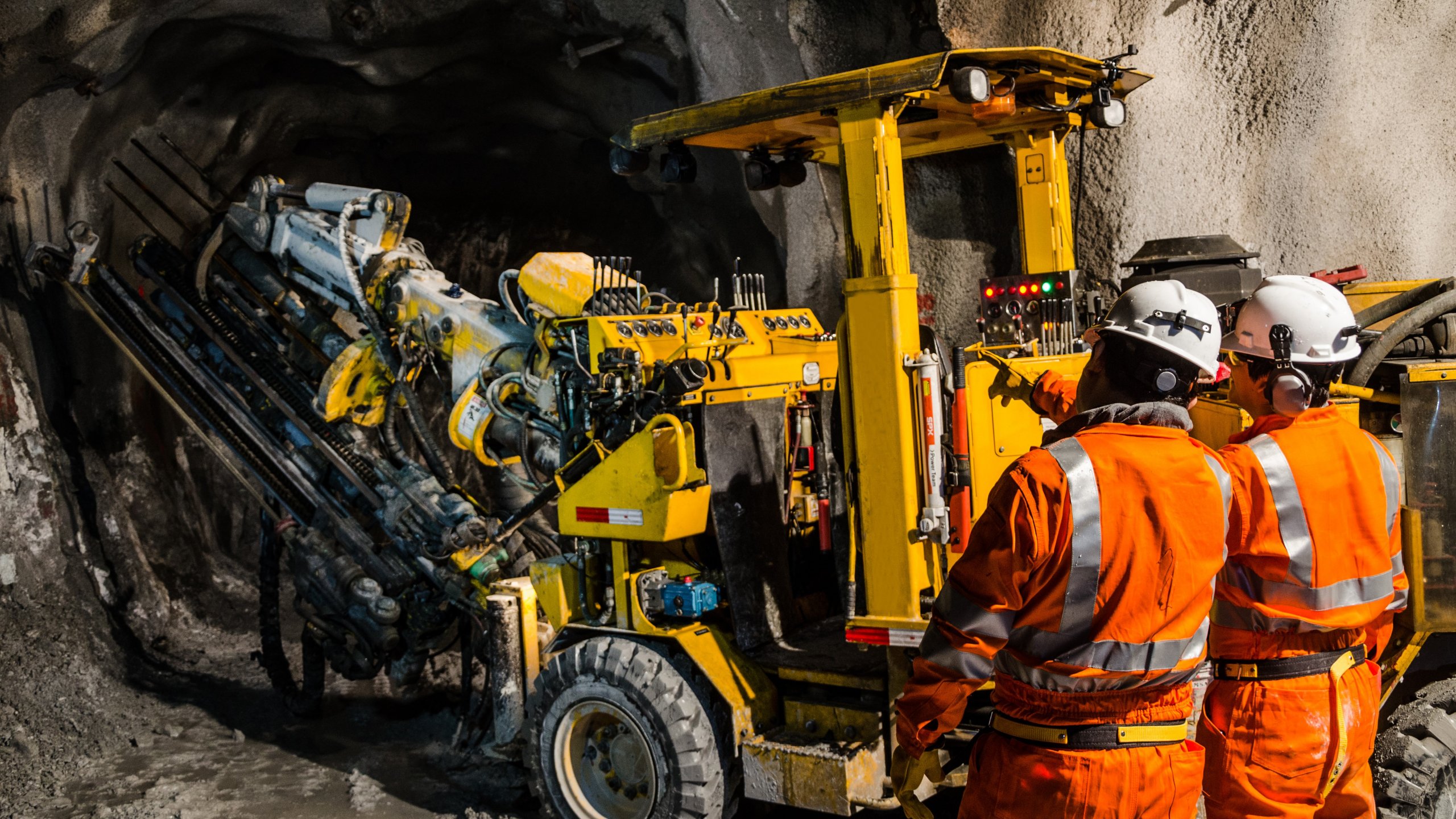 Two male mechanics in orange jumpsuits and white hard hats stand looking at yellow mining equipment in tunnel