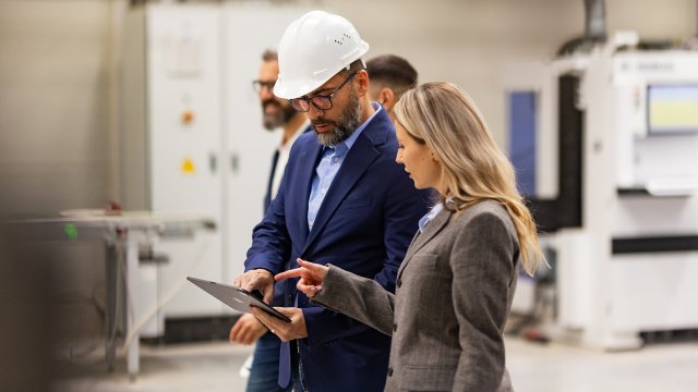 Two OT security leaders, one male and one female, standing on manufacturing plant floor holding a tablet analyzing data for NIS2 compliance.