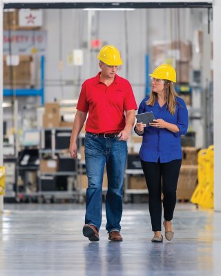Dois funcionários utilizando capacetes amarelos, caminhando e conversando em uma fábrica 