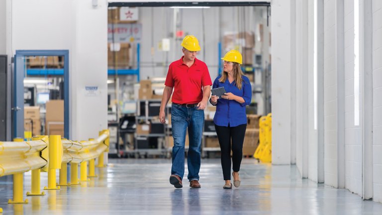 Deux employés, un homme et une femme, portant des casques de protection jaunes en pleine conversation en marchant dans une usine