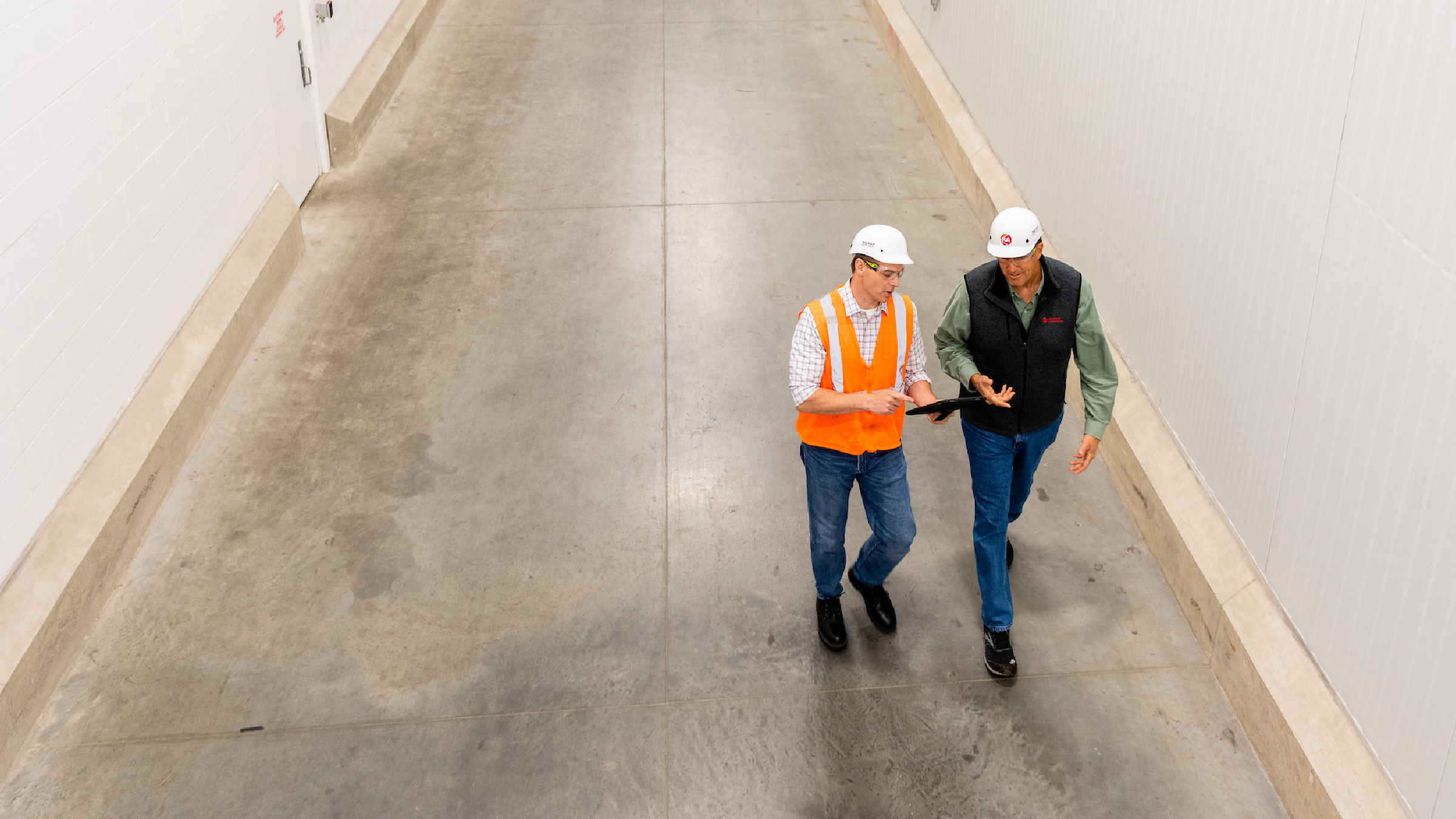 Two industrial workers wearing hard hats and safety gear walk through a clean manufacturing facility corridor while reviewing information on a digital tablet