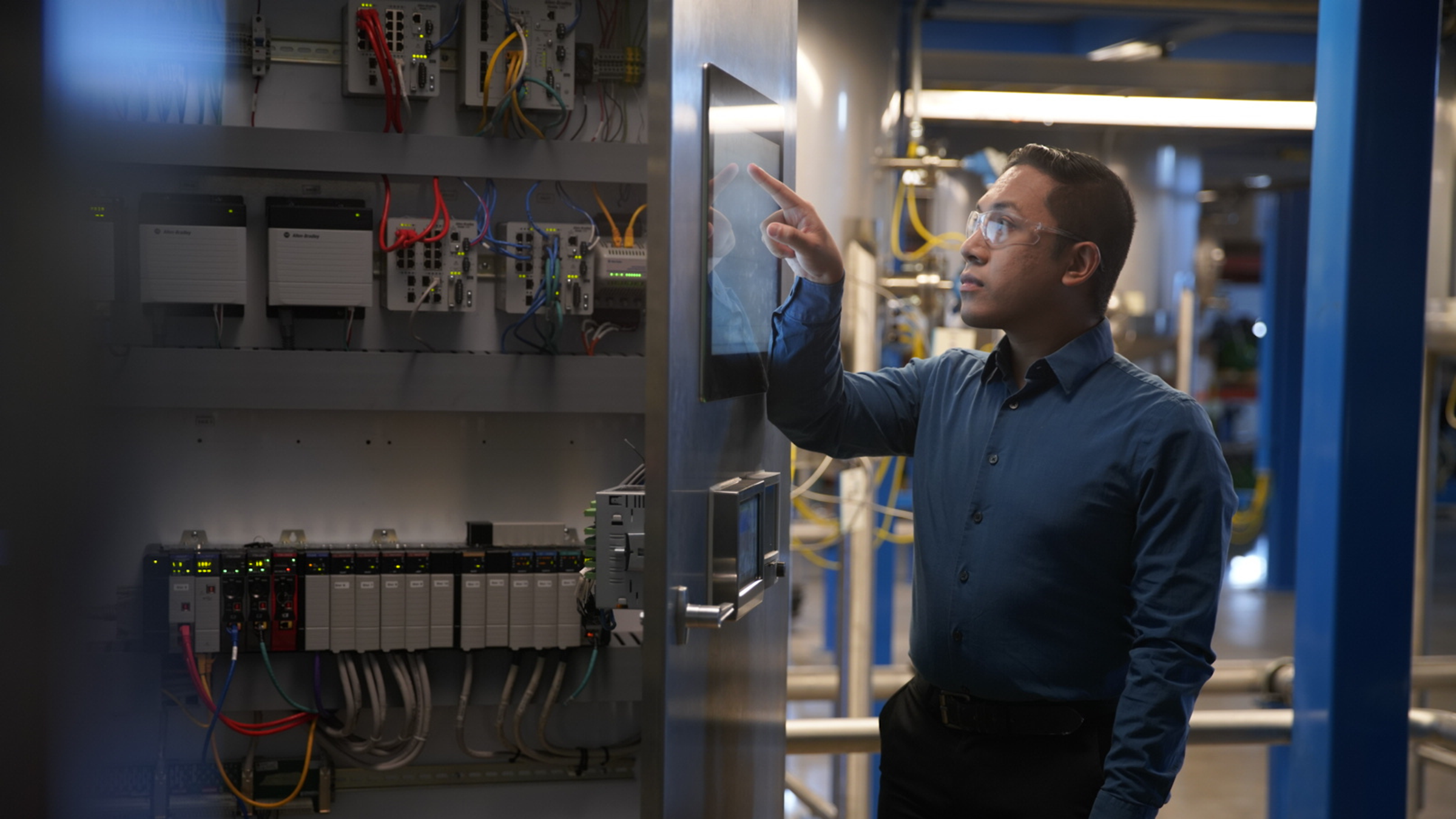 An engineer wearing safety glasses operates a touchscreen interface mounted on an industrial control cabinet door