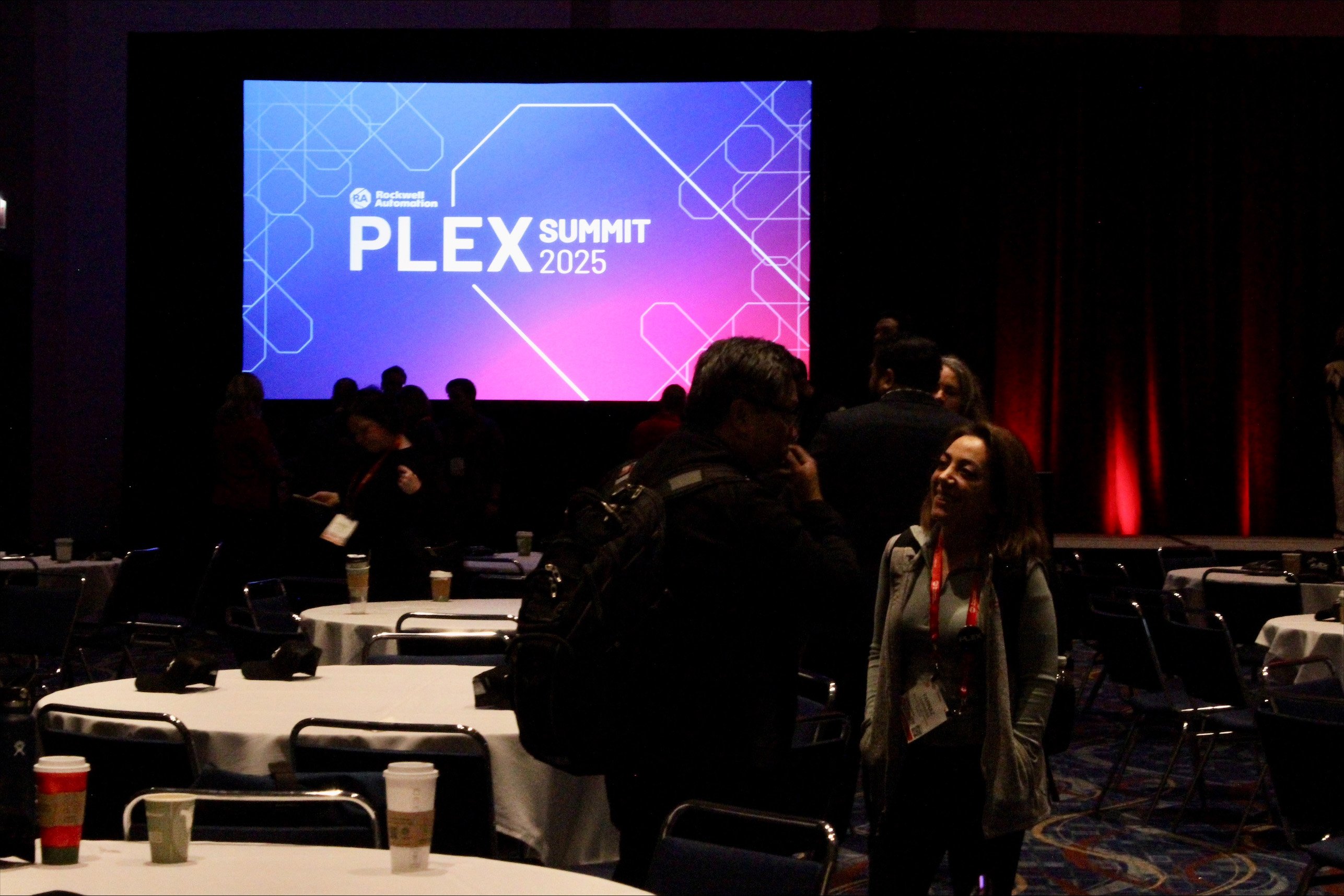 Conference room with round tables and a large screen showing ‘PLEX Summit 2025’ in blue and pink gradient.
