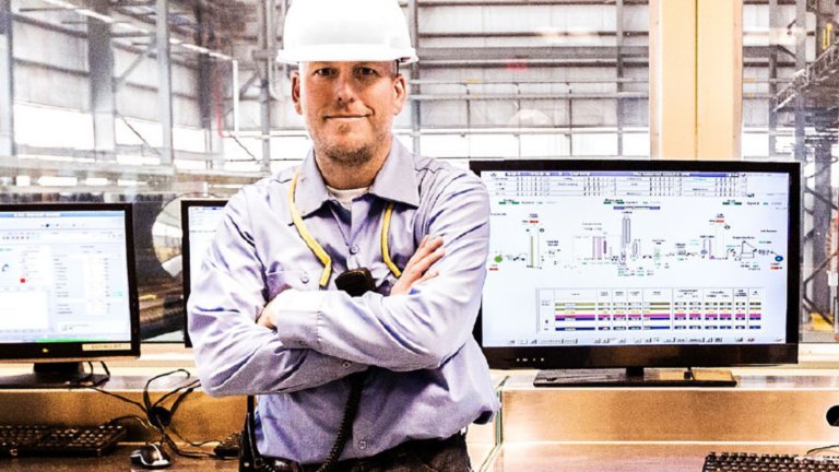 Male industrial worker wearing a hardhat with arms crossed while leaning back against a desk that has a monitor with an automation security screen in the background