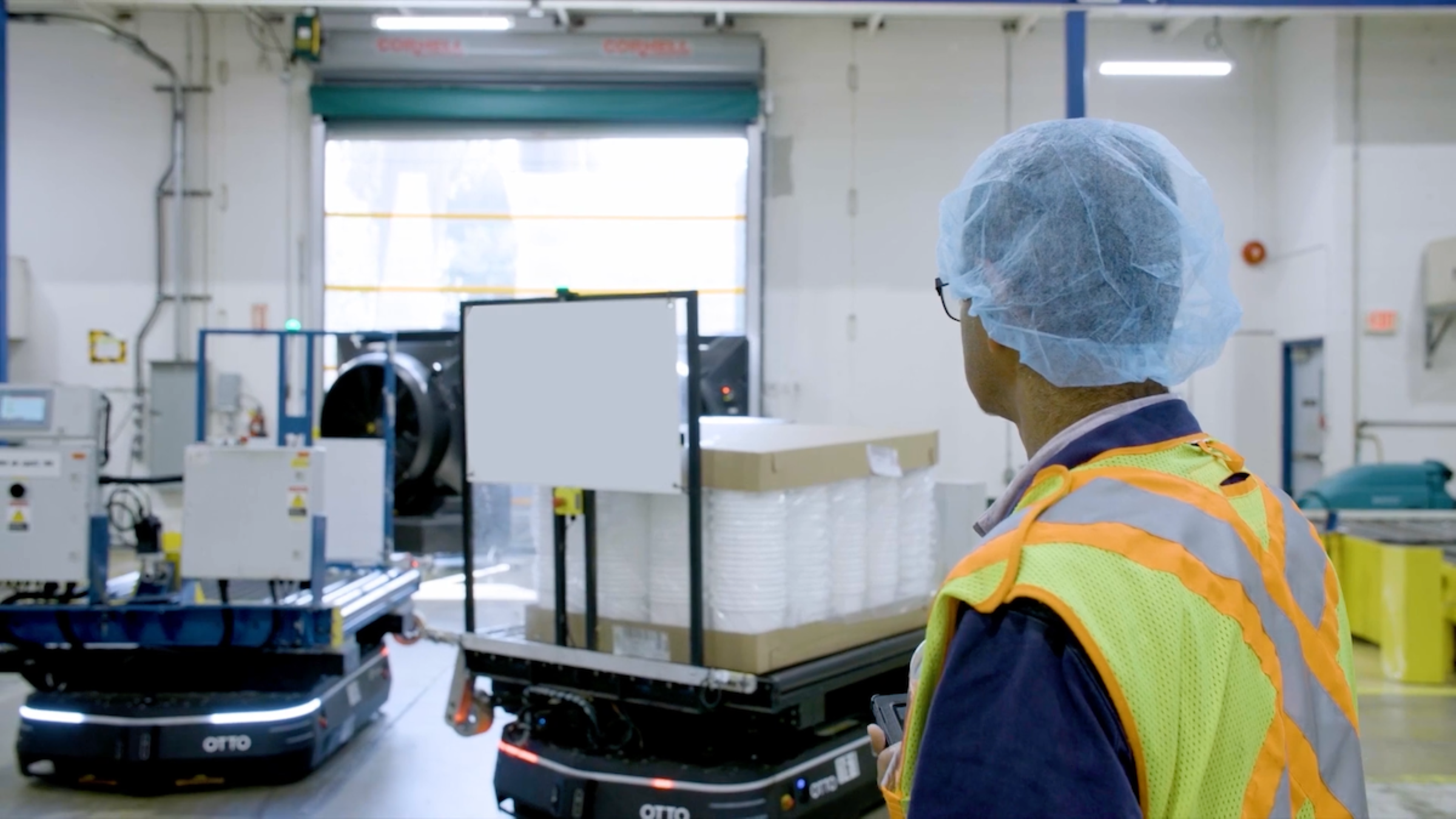 A man in a high visibility vest and a hair net looks at two OTTO 1500 AMRs operating near a dock door.