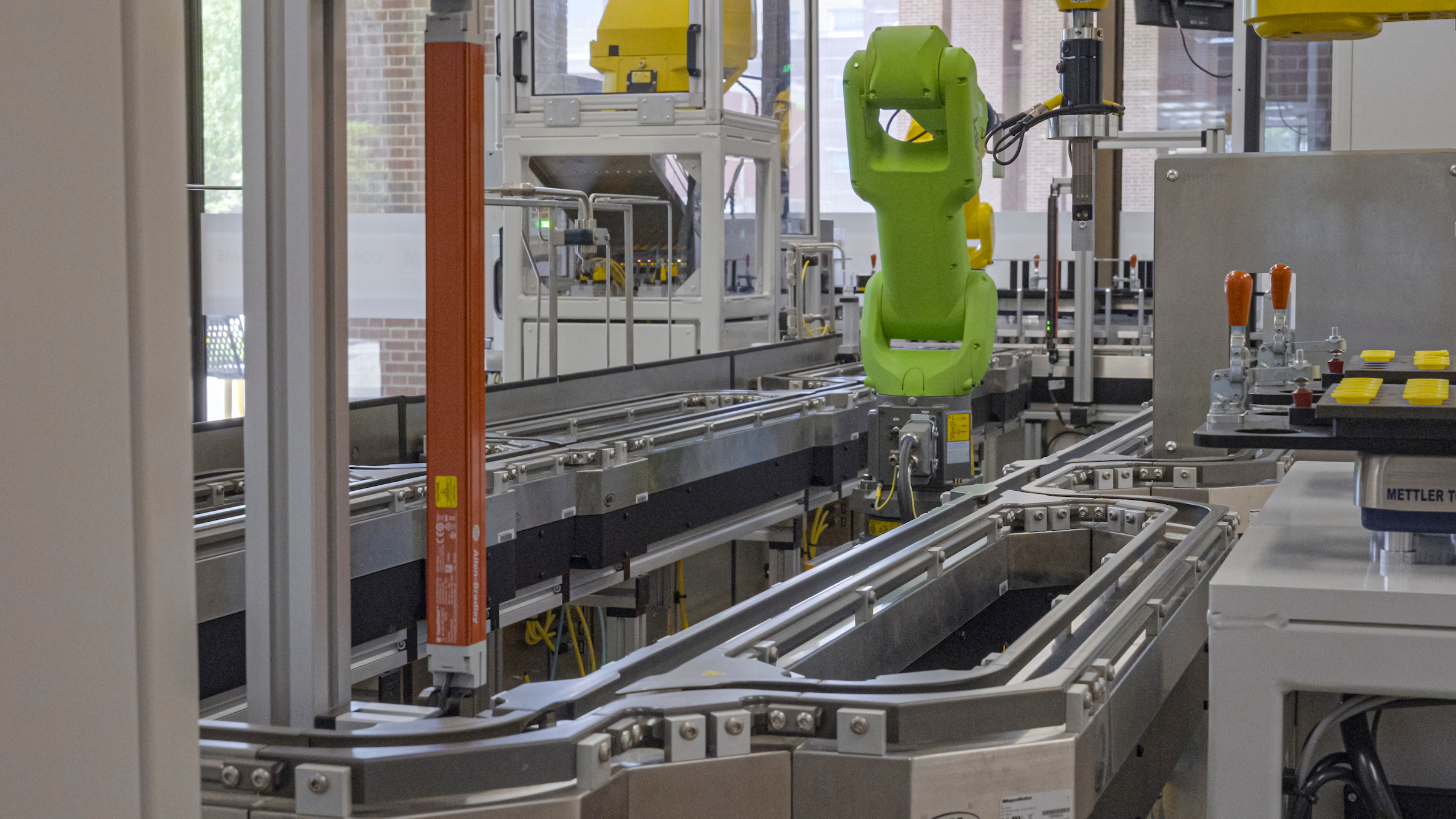 Automated production line with a green industrial robot arm operating along a conveyor system, surrounded by metal frameworks, control equipment, and packaging components in a factory environment