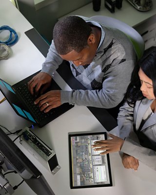 Deux employés assis sur un bureau examinent diligemment les données provenant d’un logiciel IHM sur une tablette et saisissent les résultats dans une console de programmation portable