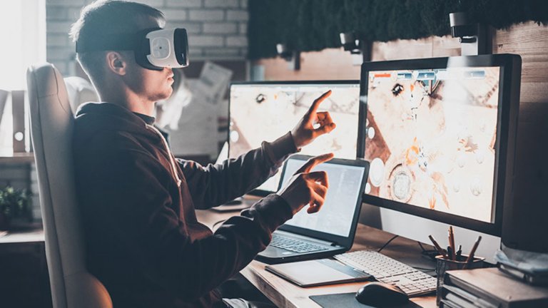Funcionário sentado em sua estação de trabalho em frente a dois monitores e de seu laptop, usando um capacete de realidade virtual