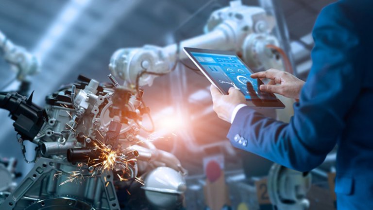The arms of a man in a blue coveralls holding a tablet in front of a welding robotic arm