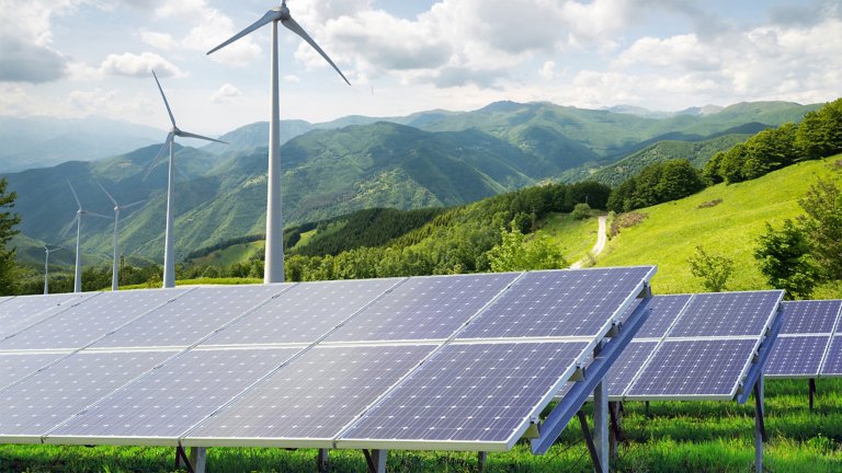 Solar panels with wind turbines against a mountain landscape against blue sky with clouds.