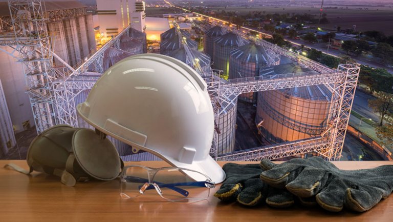 Un casco blanco, unos guantes de trabajo y unas orejeras industriales están expuestos en un escritorio con vistas a una instalación de petróleo y gas.