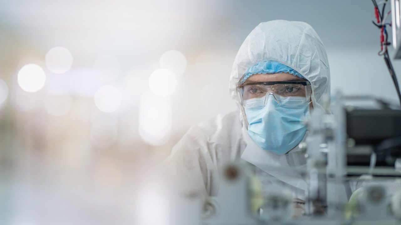 兩位生物科技專業人員穿戴全套裝備、口罩及護目鏡，在明亮的廠房中操作藥品生產設備。
