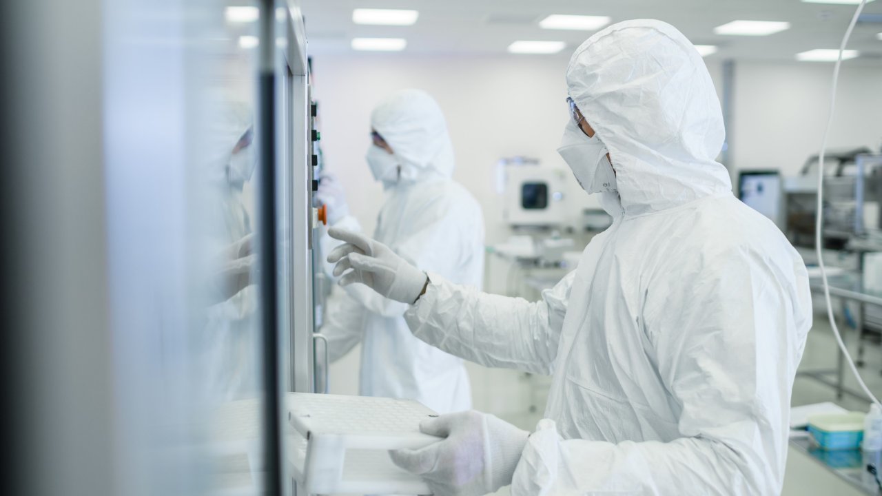 一名穿着工作服、戴着口罩和护目镜的生命科学专业人员正在对车间内的自动化制药生产线进行检查。
