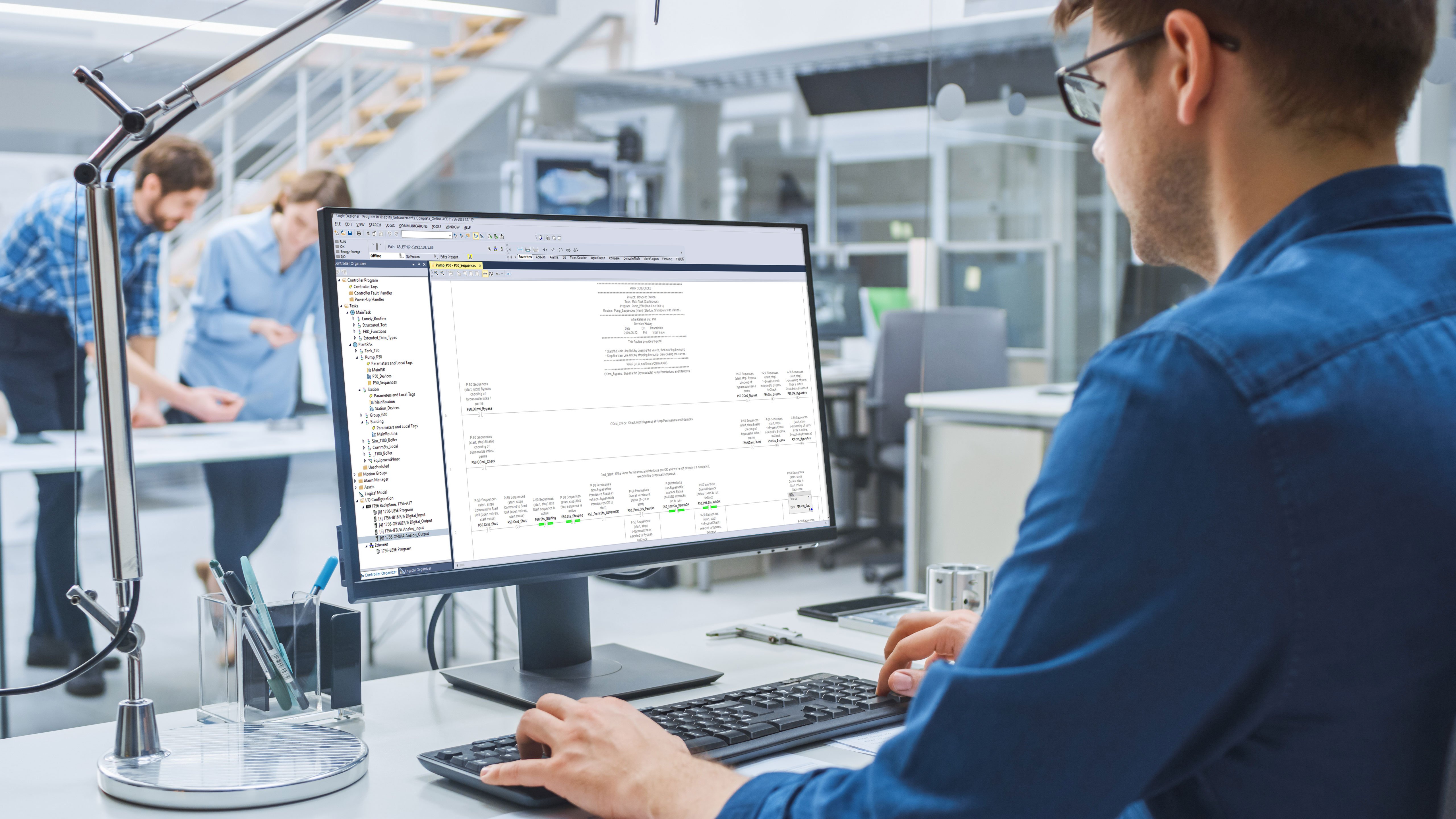 Male employee wearing glasses and entering information into a software application at his desk with two employees who are standing across from him in a deep conversation