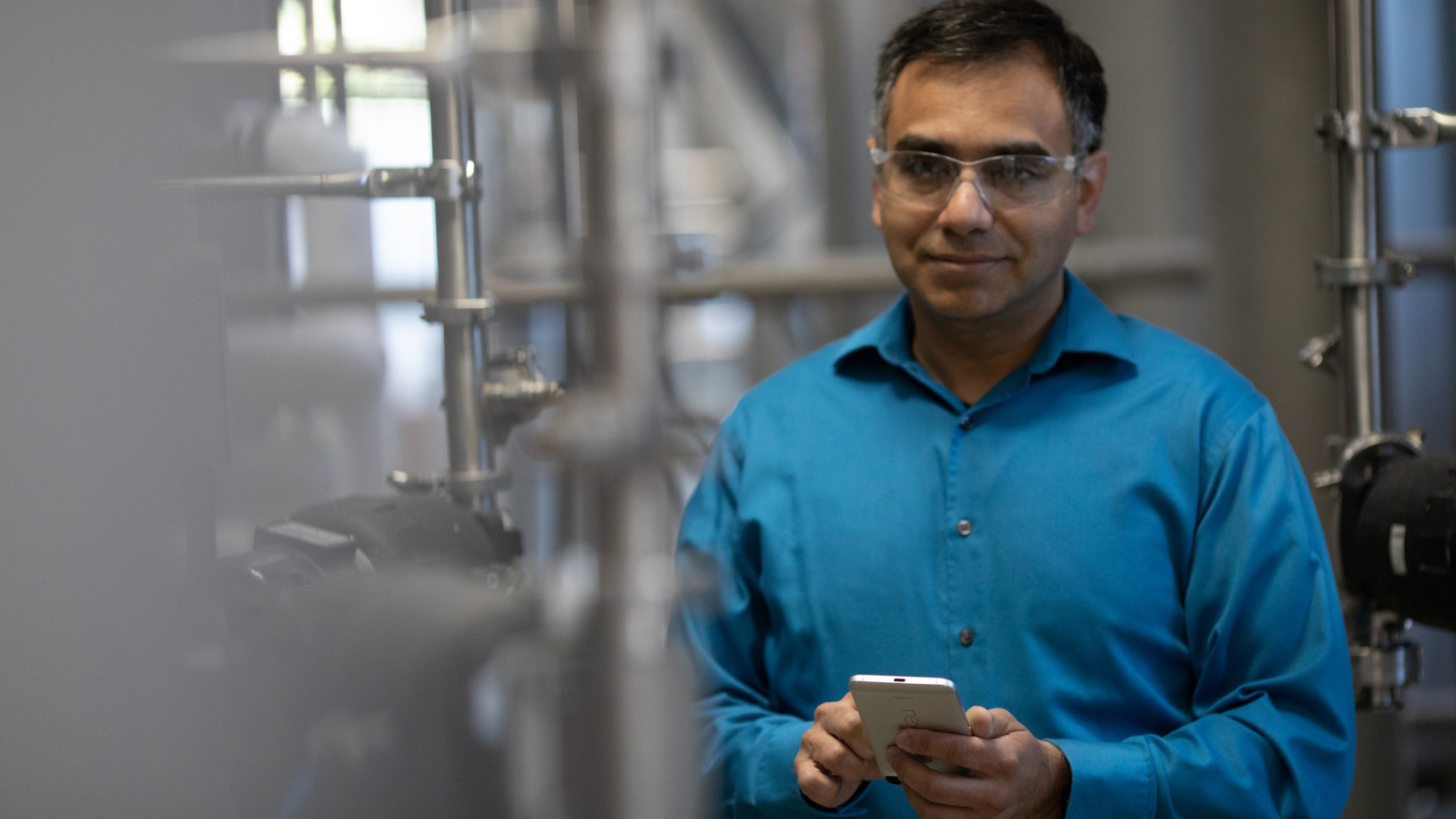 Engineer wearing a blue shirt and safety glasses in a factory looking forward holding his mobile phone