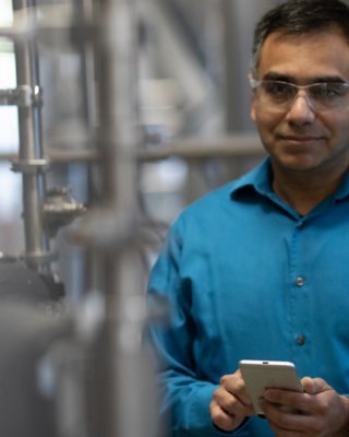 Engenheiro usando uma camisa azul e óculos de segurança em uma fábrica, olhando para frente e segurando seu telefone celular