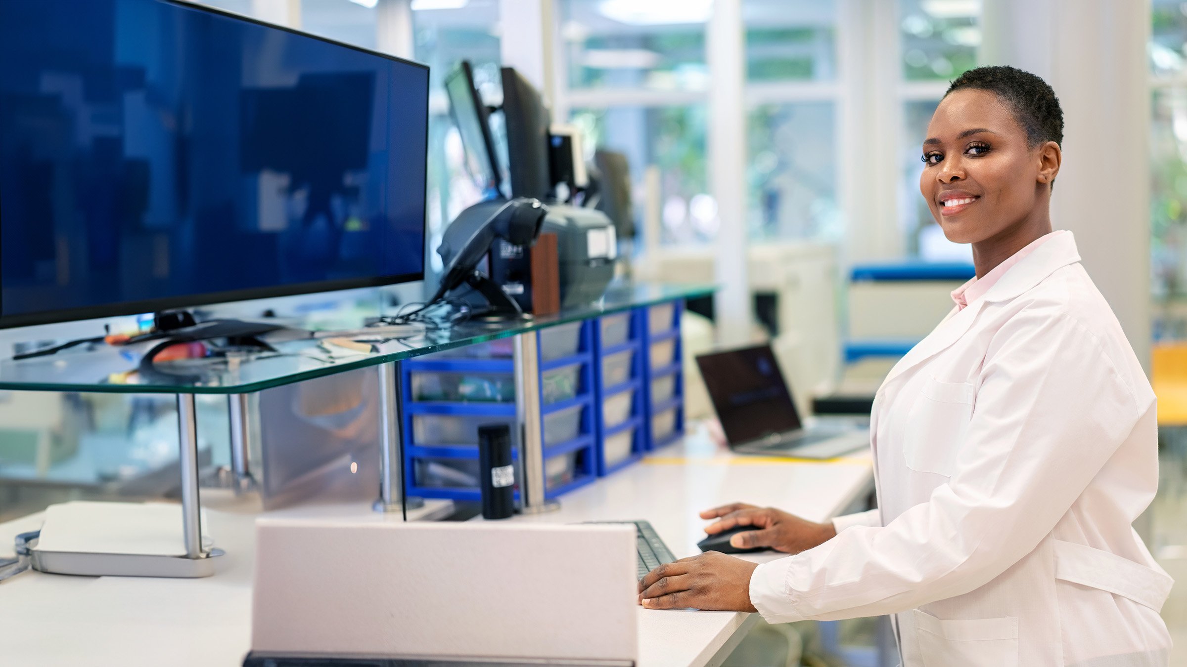 Portrait of a woman scientist working on a computer while standing at the table in a science lab and looking at camera. Female researcher wearing a lab coat working in a laboratory.