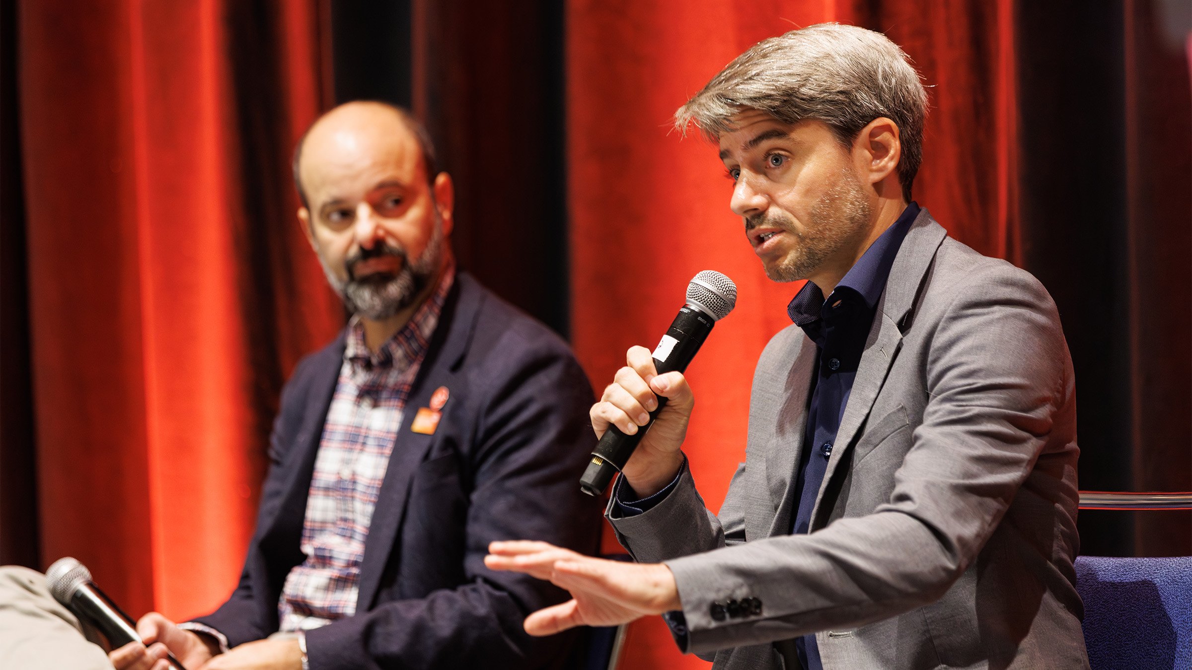 Two men on stage, one speaking with a microphone