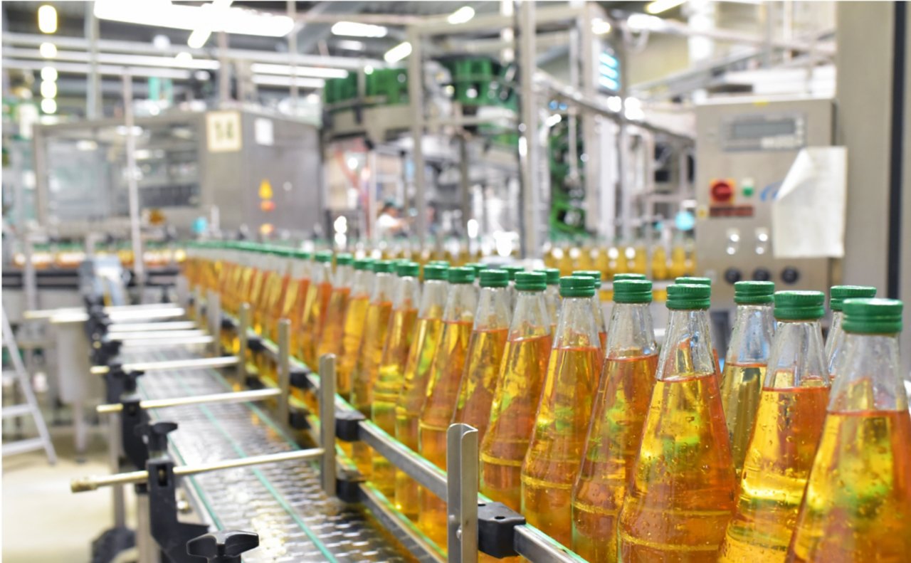 Close-up view of apple juice in clear glass bottles with green caps in a bottling factory for the food industry. Left to right, the filled bottles are on a conveyor zoomed out with factory background to zoomed in right corner foreground perspective.