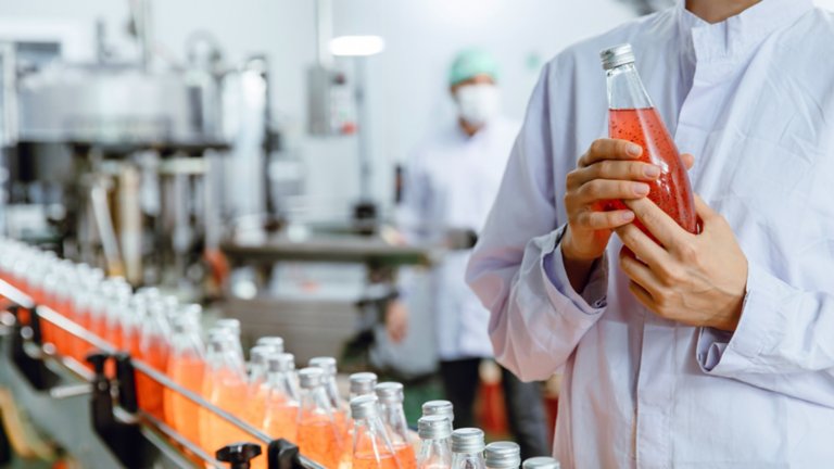 person in lab coat holding glass soda bottle by production line