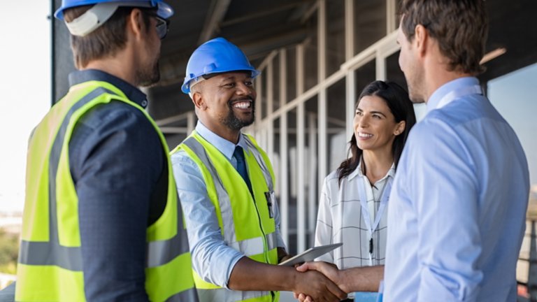 Smiling engineer shaking hands at construction site with happy architect. Handshake between cheerful african construction manager with businessman at bulding site. Team of workers with architects and contractor conclude an agreement with safety uniform.