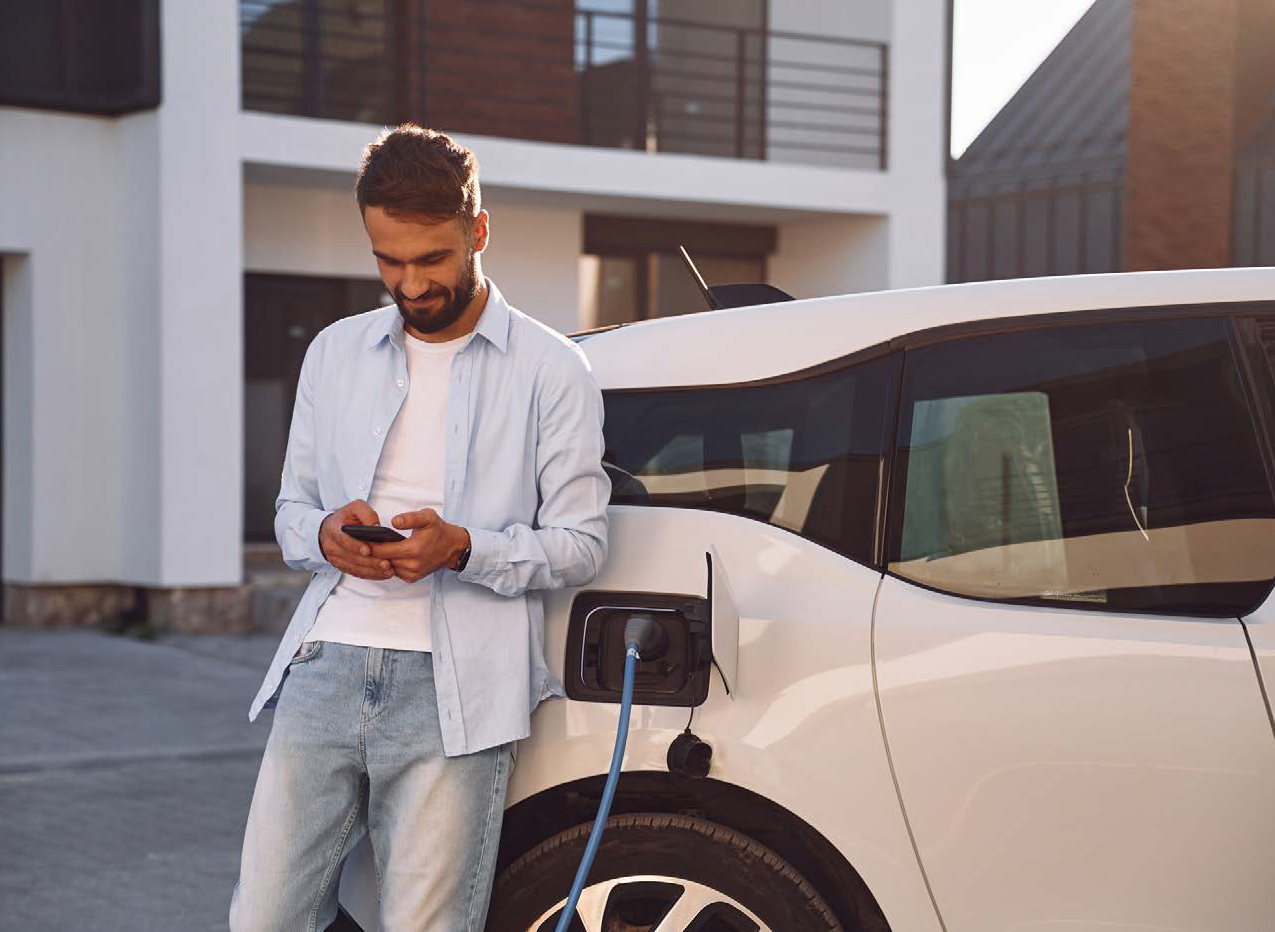 Charging the vehicle near the house. Holding smartphone. Young stylish man is with electric car at daytime.