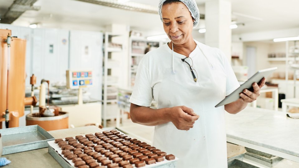 Lächelnde Arbeiterin mit einem Tablet in einer Schokoladenfabrik, die ein Blech mit frisch hergestellten Süßwaren überprüft