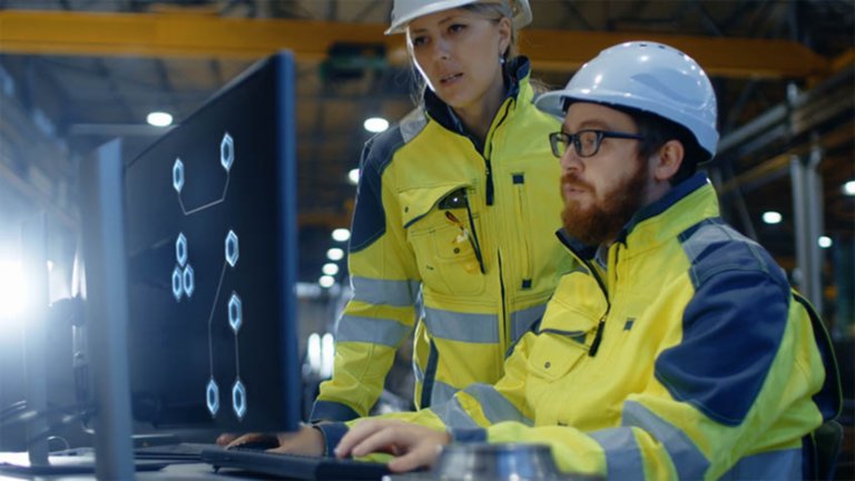 Two employees, a male and a female wearing yellow safety jackets and white hard hat looking at information on a monitor in a factory.