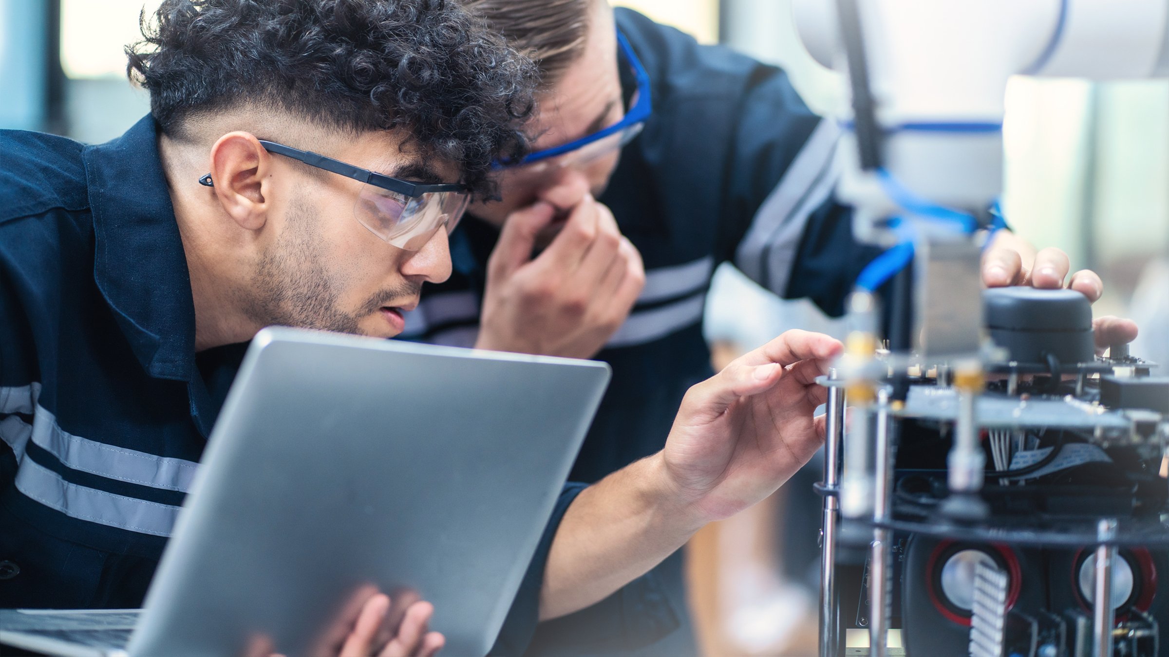 Two engineers inspect electronic and mechanical function of a hydraulic pump. One engineer is holding an open laptop.