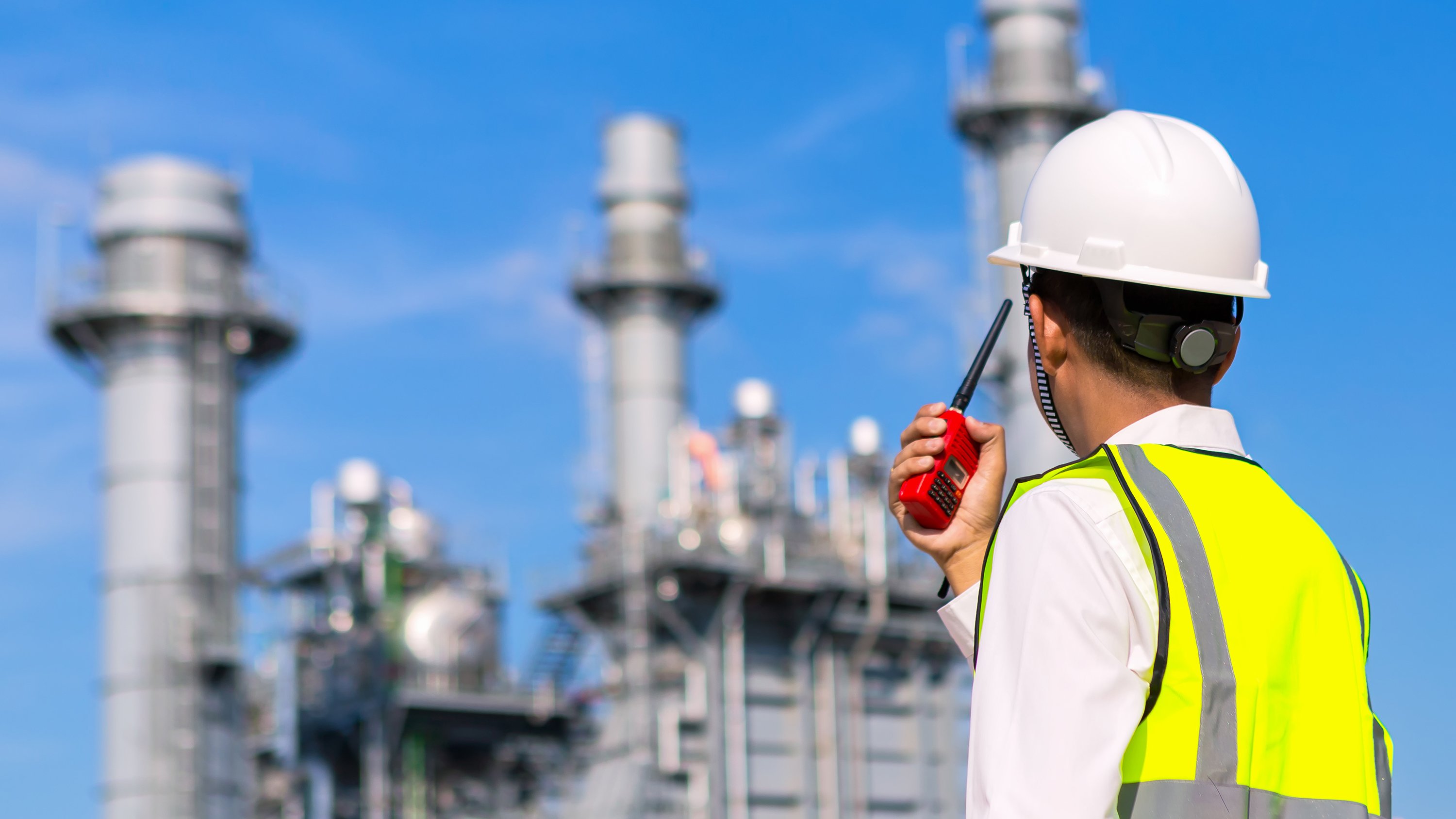 Outdoor shot of engineer in safety gear holding communication device looking up at exterior of gas plant.