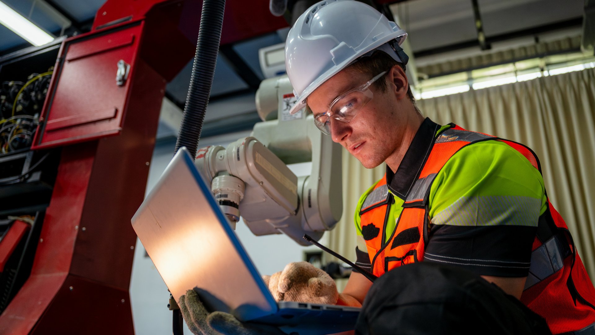 A man in a yellow and green vest is sitting on the floor and looking at a laptop. He is wearing a hard hat