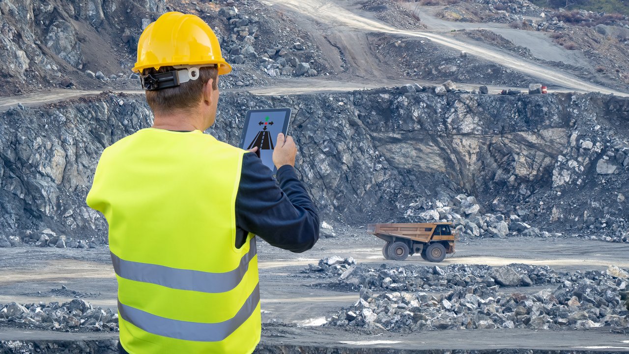 An engineer holds a tablet in his hand and remotely controls a heavy-duty dump truck in the background of a mining quarry