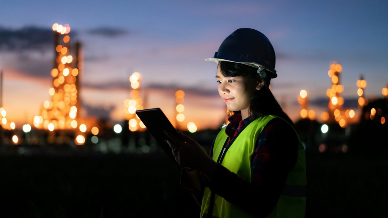 A female engineer is holding a tablet outside with a chemical plant in the distance