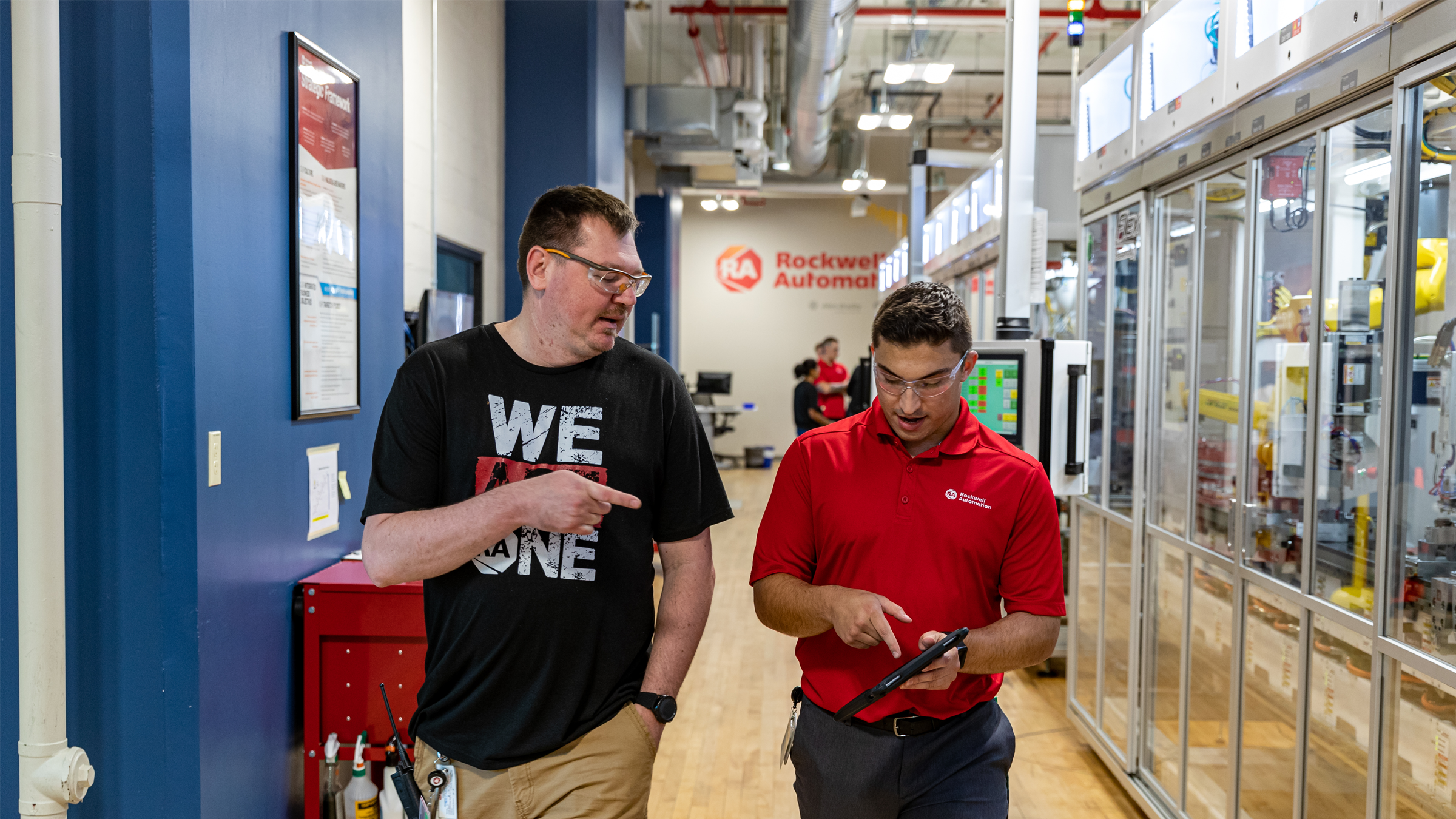 Front view of two people wearing safety glasses, one in a black shirt and one in a red shirt holding a tablet, walking by a manufacturing line