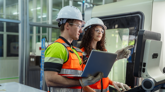 Two people wearing safety gear and holding a laptop.. Team of diverse engineers, male and female, programming and operating a CNC machine in a modern factory.
