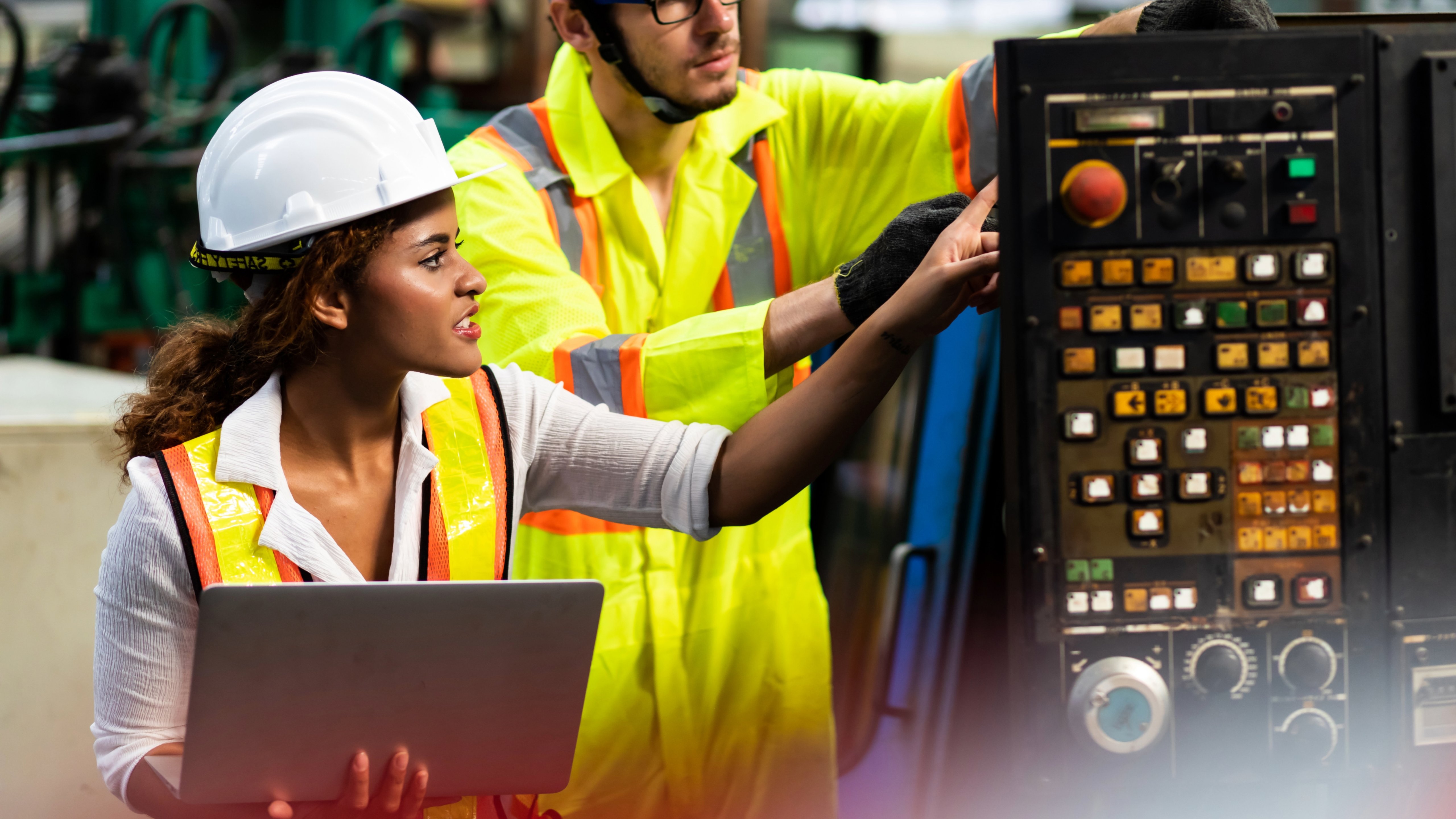 Two engineers work on a computer and a panel in a manufacturing plant.