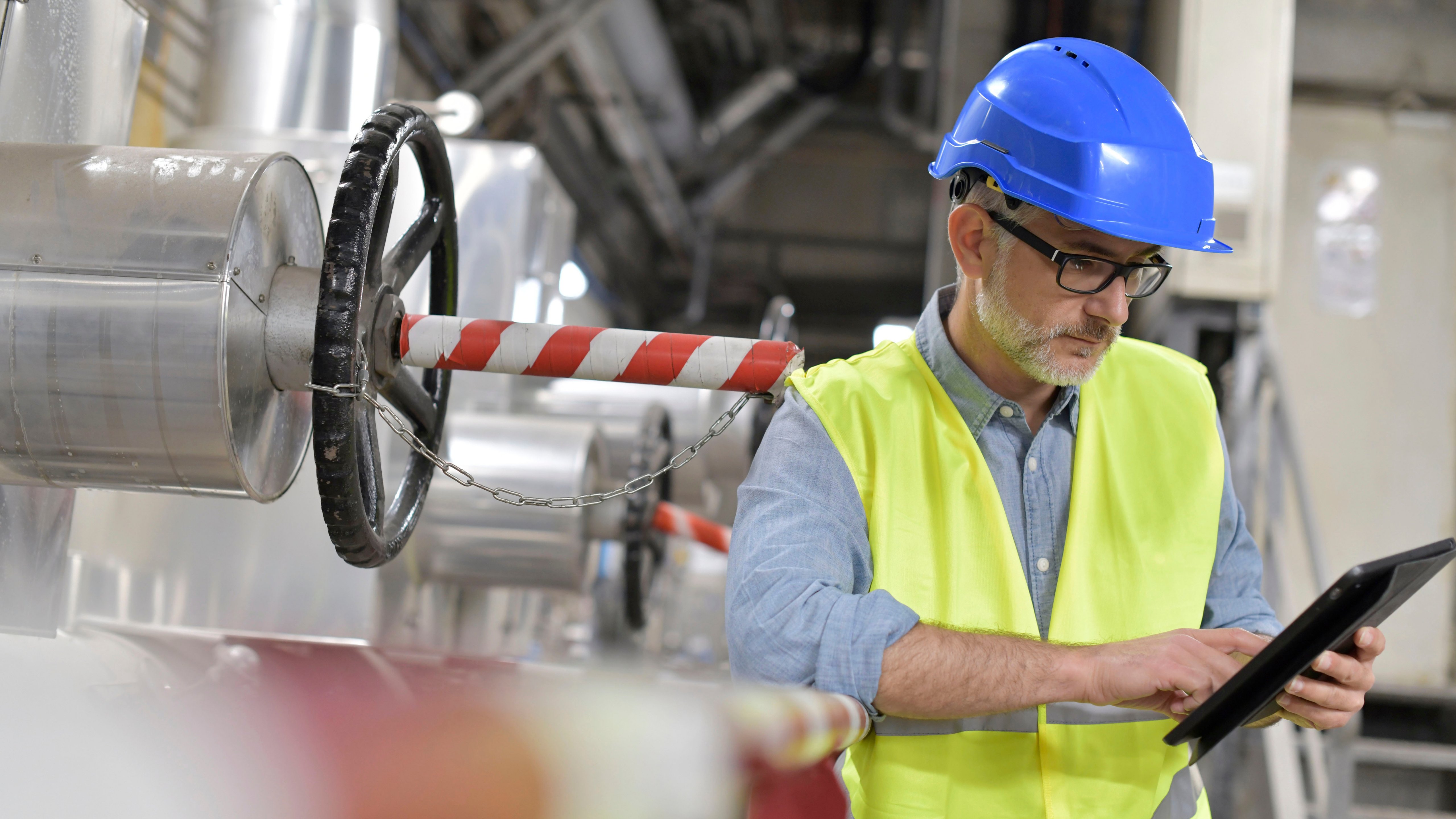 A man with a blue hard hard and reflective vest working in a factory and holding a tablet.