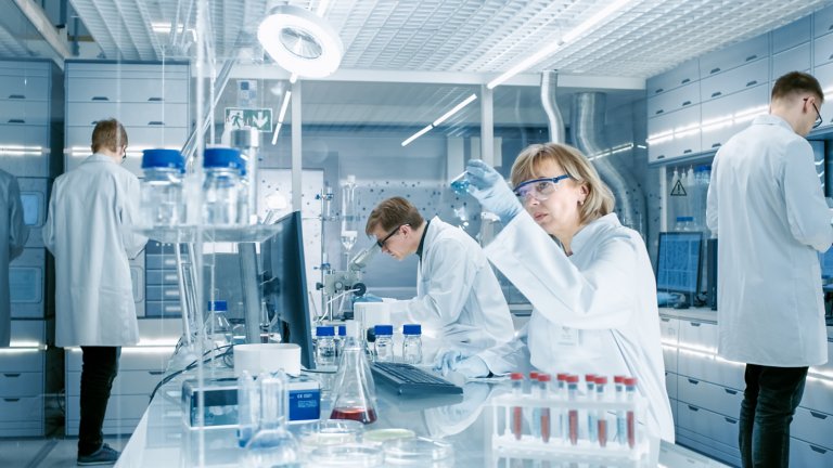 Female Scientist Analyzes Liquid in the Beaker and Types Down Observations on Her Computer. She's Working in a Busy Laboratory Full of Scientists Conducting Experiments.