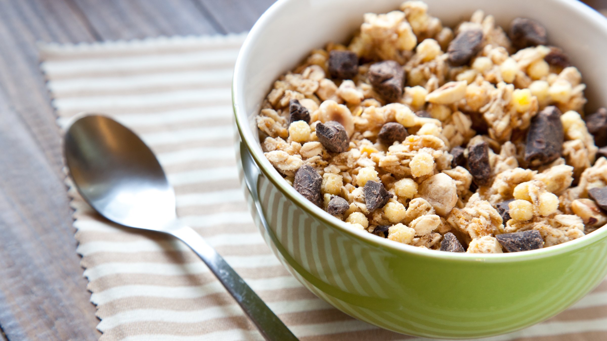 Tazón verde con granola y trozos de chocolate sobre una servilleta a rayas, con una cuchara al lado